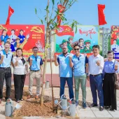 A group of nine people stand in front of a newly planted tree, smiling and making small heart gestures with their fingers. Behind them are colorful posters. Red flags wave in the background, and the setting is a sunny, paved courtyard.