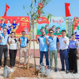 A group of nine people stand in front of a newly planted tree, smiling and making small heart gestures with their fingers. Behind them are colorful posters. Red flags wave in the background, and the setting is a sunny, paved courtyard.